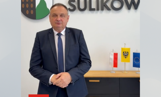 Mężczyzna w granatowym garniturze i niebieskim krawacie stoi w urzędzie na tle napisu „GMINA SULIKÓW” oraz herbu gminy. Na dole grafiki widnieje podpis: „Jan Majowski, Wójt Gminy Sulików”. Obok znajdują się małe flagi Polski, Dolnego Śląska i Unii Europej