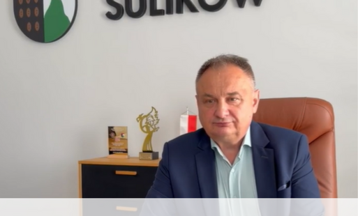 Mężczyzna w granatowym garniturze siedzi przy biurku w urzędzie gminy. Na ścianie za nim widnieje herb oraz napis „GMINA SULIKÓW”. W dolnej części grafiki znajduje się napis: „Informacja dotycząca oświetlenia ulicznego na terenie Gminy Sulików [film]”.